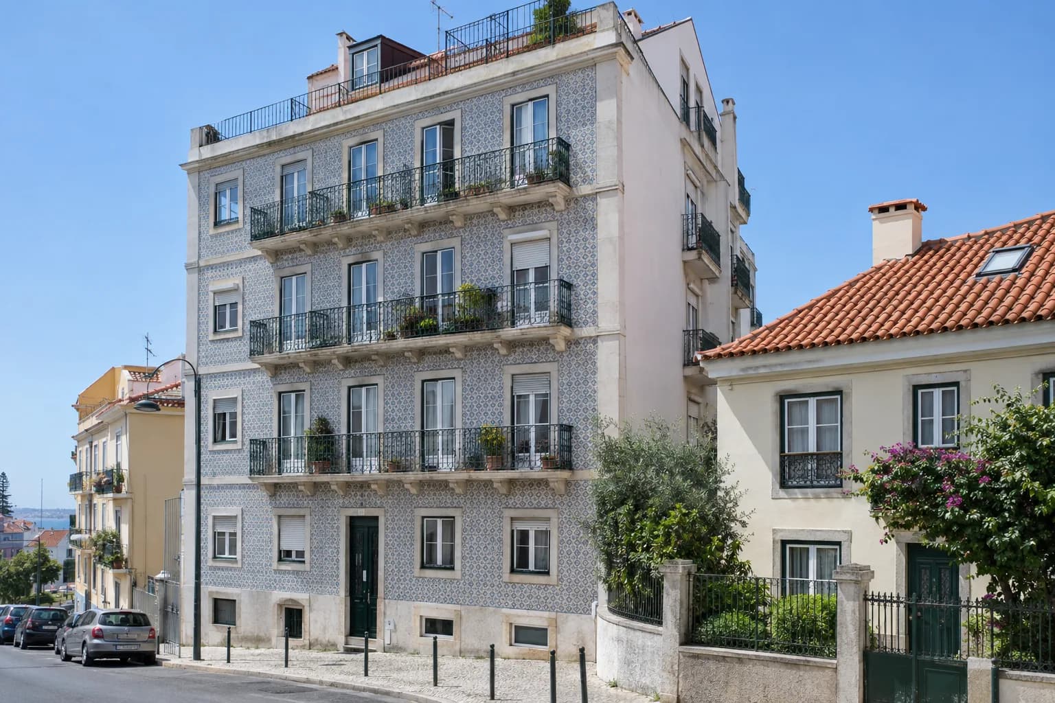 Prédio de apartamentos com fachada de azulejo e varandas de ferro numa rua de Lisboa, ao lado de uma casa com telhado de telha