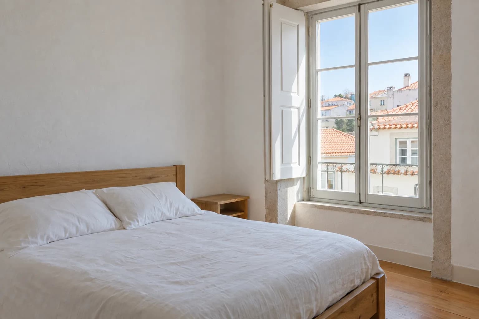 Quarto com cama de madeira e roupa de cama branca, junto a uma janela com portadas que dá para telhados de telha