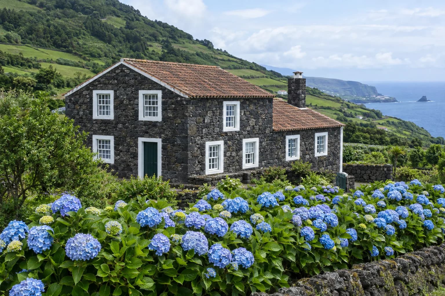 Casa de pedra basáltica escura, com molduras brancas nas janelas e telhado de telha, rodeada de hortênsias numa encosta junto ao mar