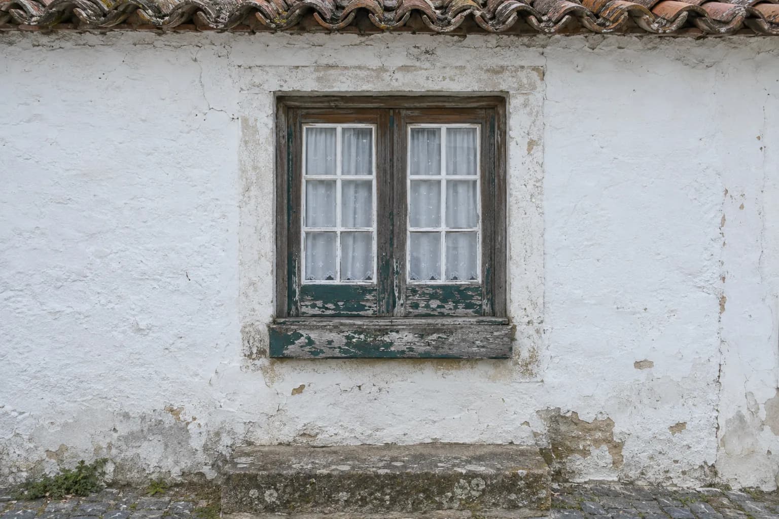 Janela de madeira com a pintura a descascar, numa parede caiada de uma casa antiga, com beirado de telha por cima